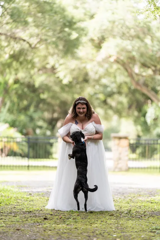Samantha in a white dress holds a black dog upright in her arms while standing on the lawn.