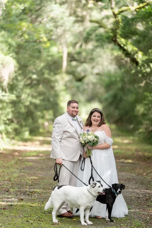 The couple stands on a shaded path holding leashes of a black and white dog while smiling at each other.