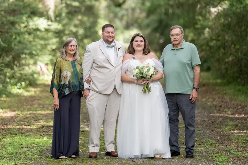 Matthew and Samantha pose with two family members beneath towering trees, Samantha holding her bouquet.