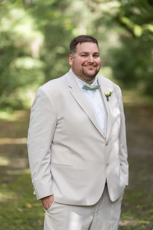 Matthew smiles in his cream suit with a floral boutonniere, oak trees softly blurred behind him.