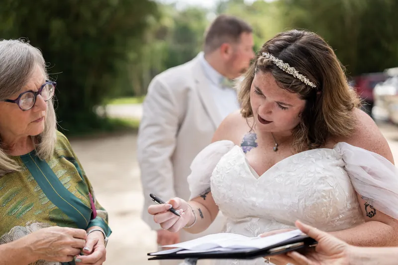 Samantha signs a document while a woman in glasses looks on and Matthew stands nearby at the park.