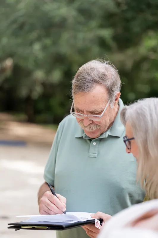 An officiant in a light blue polo and glasses watches as someone signs paperwork during an outdoor ceremony.