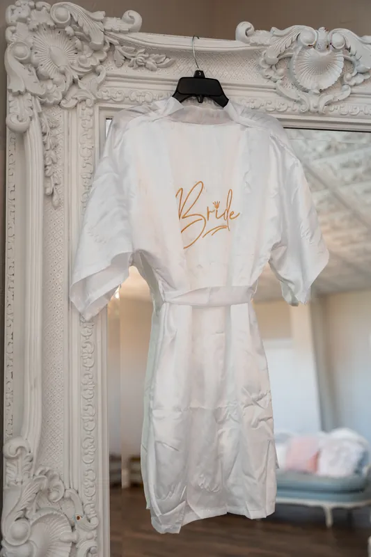White silk bridal robe with "Bride" embroidered in gold hanging on an ornate white mirror frame at The White Room, St.