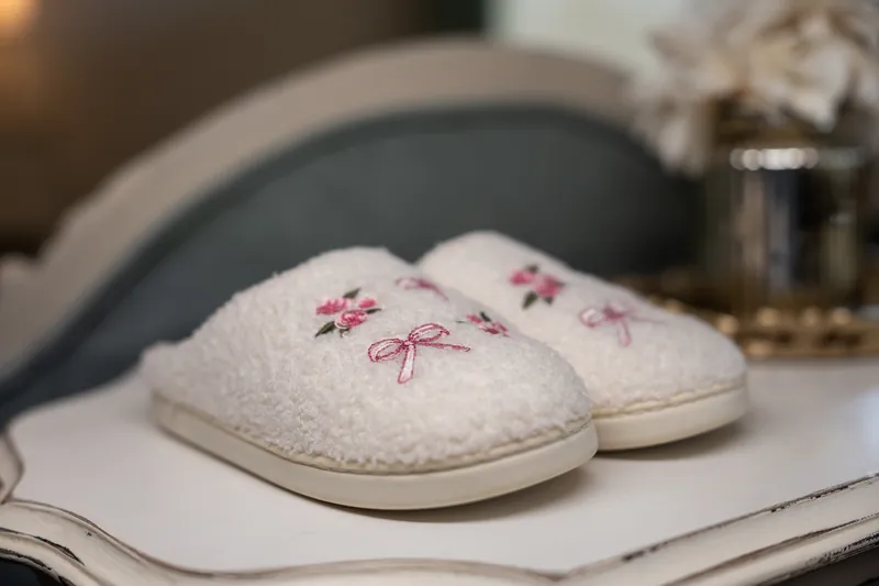 Pair of cream slippers embroidered with pink bows rest on a white surface near a gray chair and bedside table.