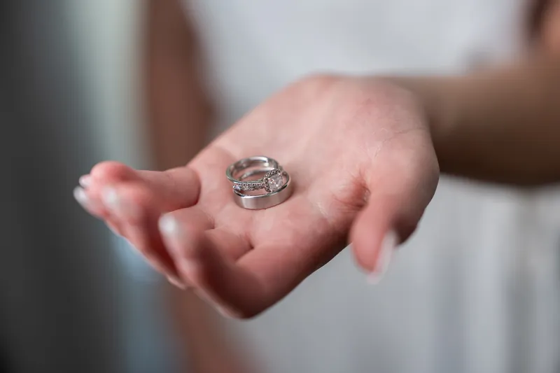 Sarah holds her diamond engagement and wedding rings in an open palm, wearing a white dress.