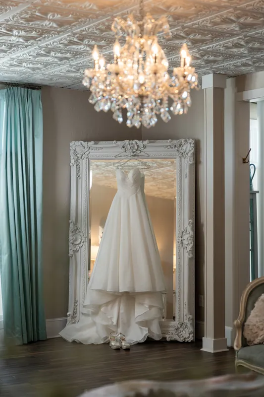 Sarah's flowing white wedding dress hangs in an ornate silver-framed mirror beneath a crystal chandelier at The White Room