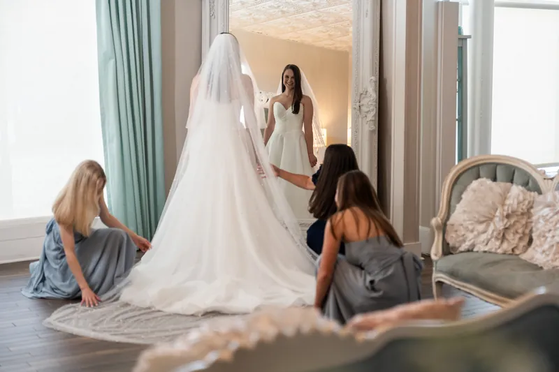 Bridesmaids arrange Sarah's train while she stands in her white gown near floor-to-ceiling windows.