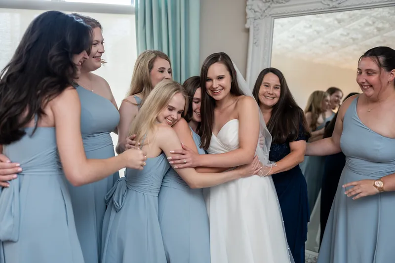 Sarah laughs surrounded by bridesmaids in pale blue dresses during getting-ready moments.