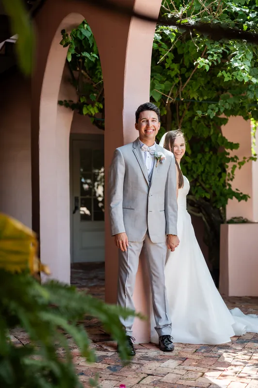 Nathan stands in a light blue suit beneath ivy-covered arches at The White Room, holding Sarah's flowing white train.