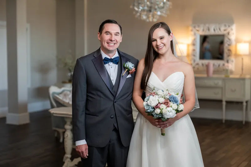 Nathan and Sarah stand together in formal attire, he in black tuxedo and she in her strapless gown holding flowers.