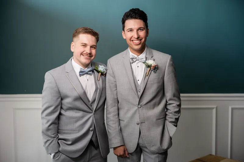 Nathan and his groomsman stand shoulder-to-shoulder in matching gray suits against a teal and white wall at The White Room.