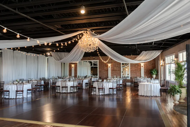 Elegant reception hall draped with flowing white fabric, string lights, and dark wood dance floor lined with guest tables.