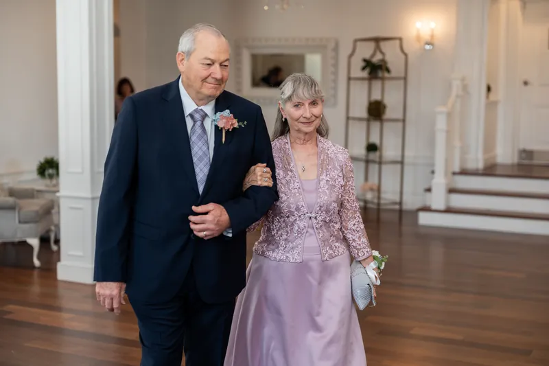 Sarah's father escorts her down the aisle in a navy suit while Sarah carries her white bouquet in a lavender gown.
