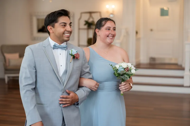 Sarah and Nathan walk arm-in-arm through the venue, both beaming, with Sarah in her blue gown and holding a white bouquet.
