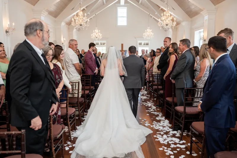 Sarah processes down the aisle at The White Room as guests stand and watch, her long train trailing behind on the wooden