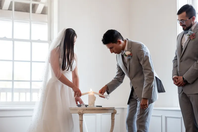 Sarah and Nathan exchange vows at a wooden table while guests and groomsmen watch in The White Room's bright, windowed