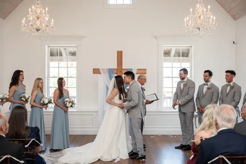 Sarah looks at Nathan as he speaks during their vows, with bridesmaids and groomsmen flanking them in the white-walled venue.