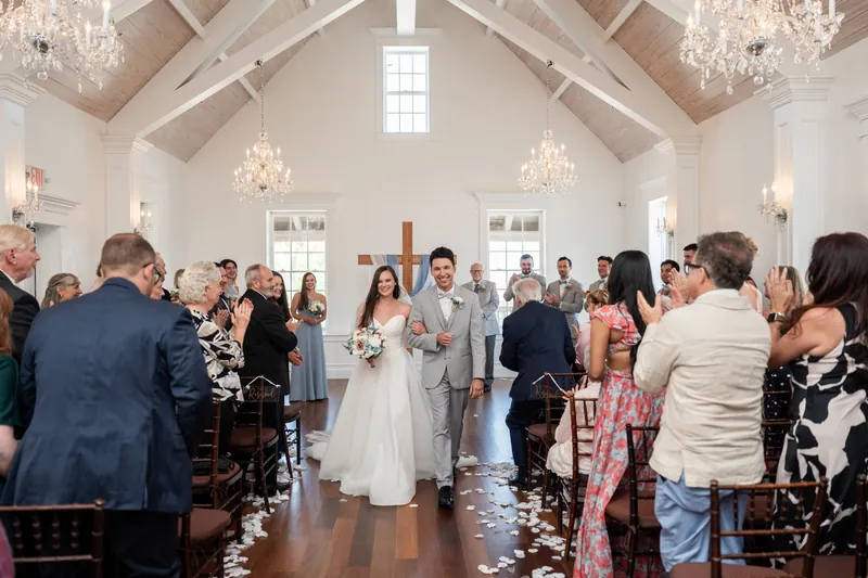 Guests rise from wooden chairs to celebrate as Sarah and Nathan kiss at the altar, confetti scattered on the aisle floor.