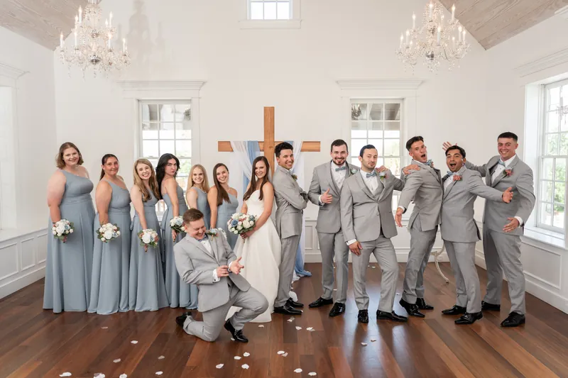 Sarah stands centered in her white gown surrounded by bridesmaids in dusty blue dresses and groomsmen in gray suits inside