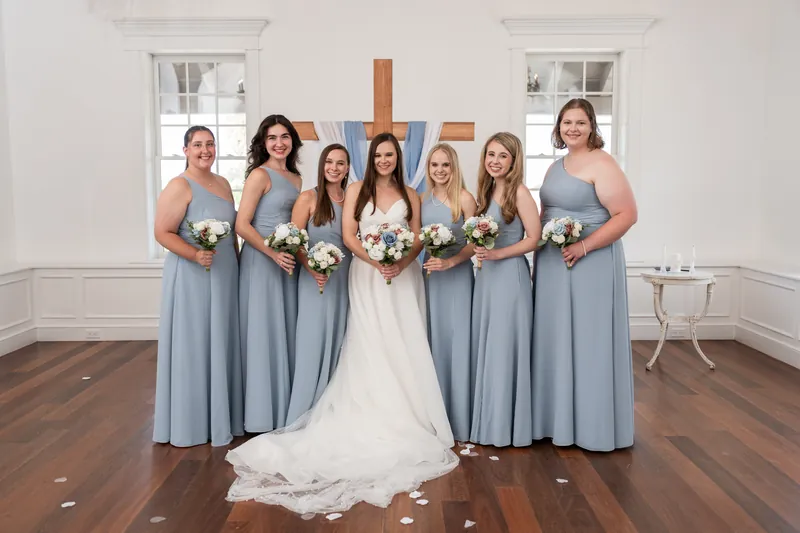 Sarah poses with seven bridesmaids in matching dusty blue dresses, all holding white bouquets on the wooden floor