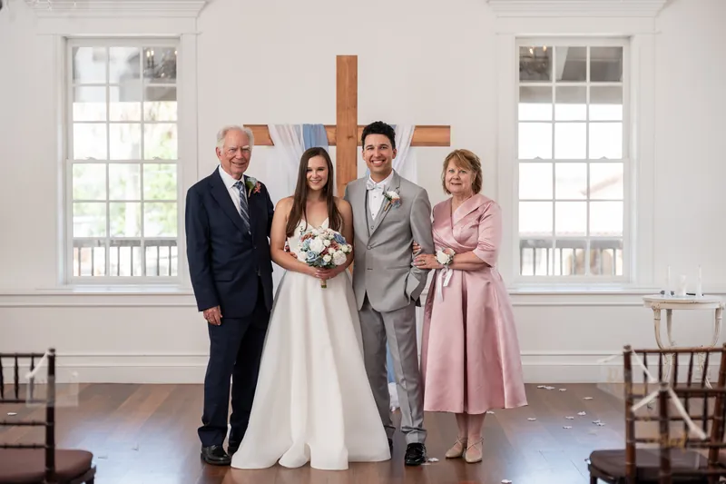 Sarah holds her bouquet between her father in a dark suit and Nathan in gray, with his mother in a pink dress beside them