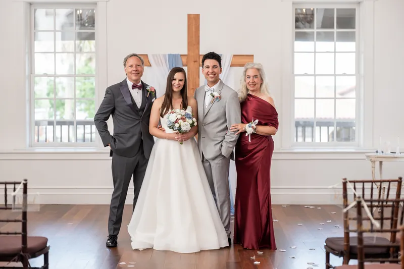 Sarah and Nathan pose with two family members—a man in gray and a woman in burgundy—in the bright ceremonial hall