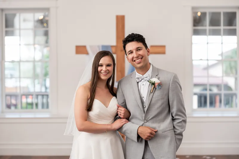 Sarah and Nathan stand together before a wooden cross at The White Room, both smiling in their wedding attire.