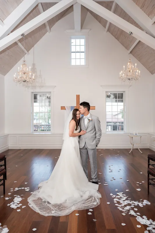 Nathan kisses Sarah's forehead as her lace train pools around them on the wooden floor beneath vaulted ceilings.