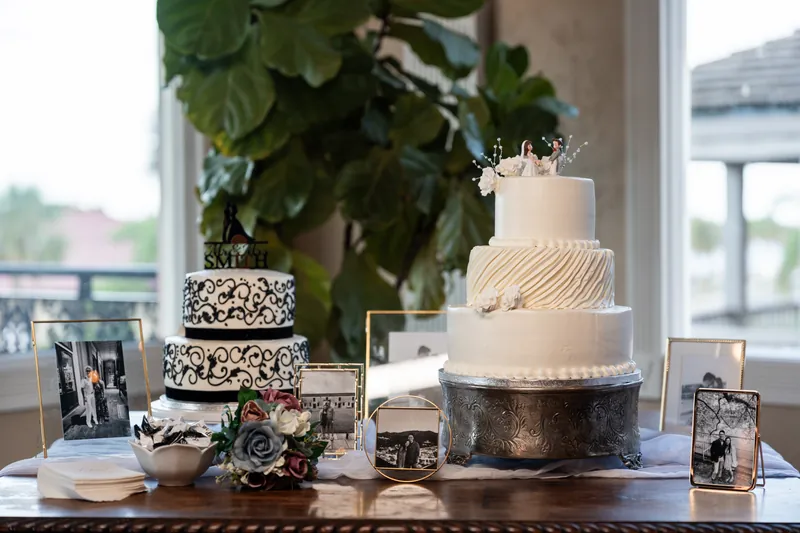 A three-tiered wedding cake with white and patterned details sits on display beside greenery and framed photos.
