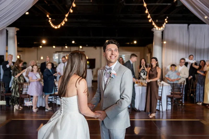 Sarah and Nathan hold hands during their first dance as guests watch from around the wooden floor.