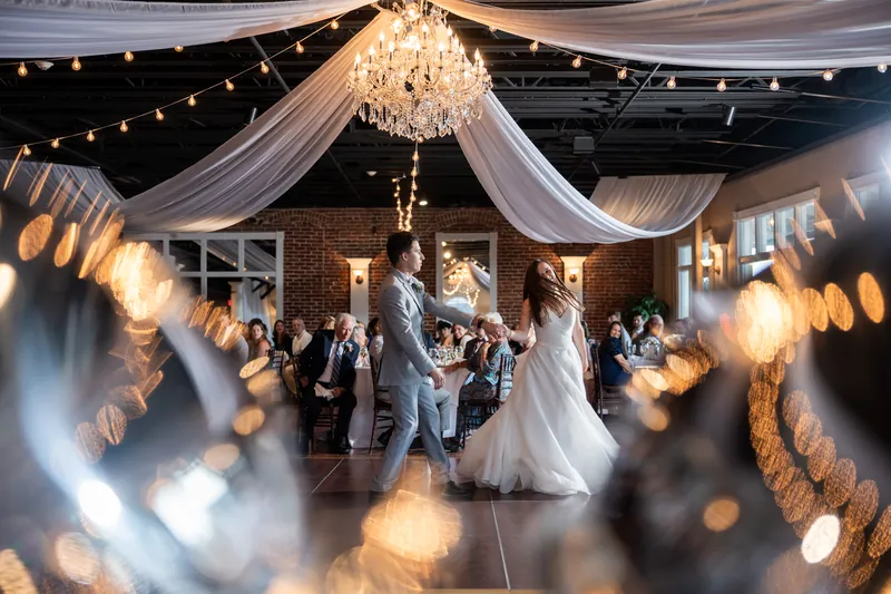Warm string lights and draped fabric frame the couple dancing amid glowing bokeh and guests celebrating around them.