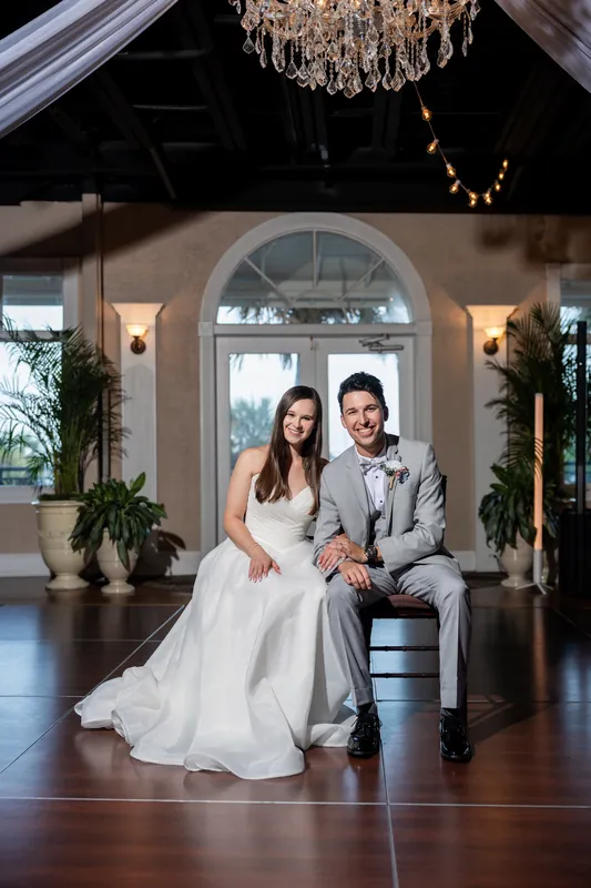Sarah and Nathan sit together on a bench beneath a crystal chandelier and arched doorway at The White Room, surrounded