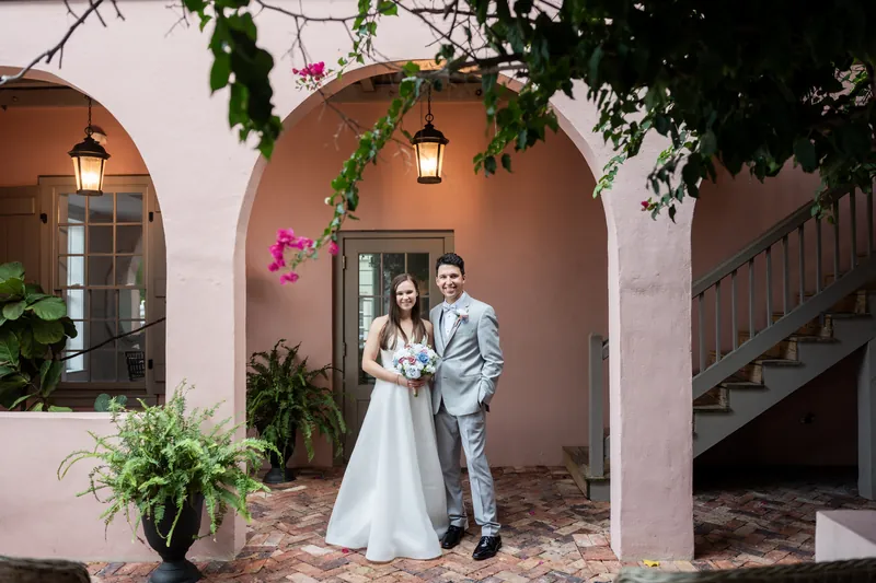 Sarah and Nathan stand hand-in-hand under a blooming magenta bougainvillea in the courtyard's Spanish Colonial archway.