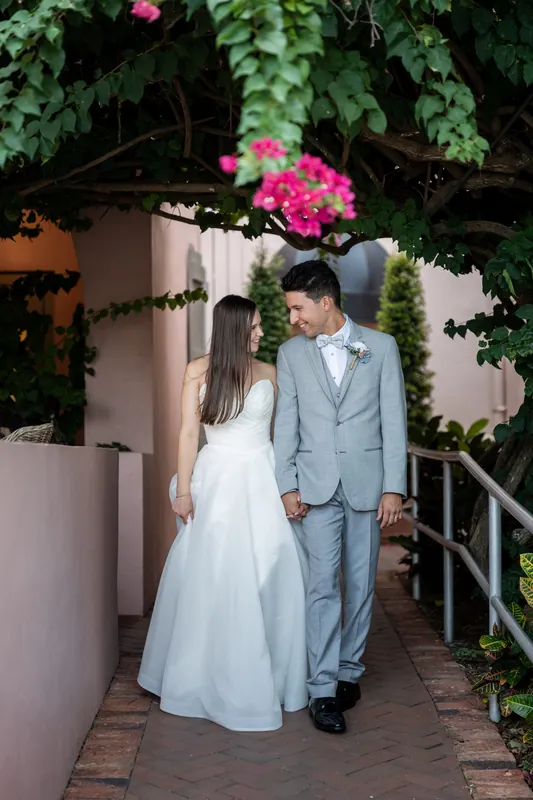 The couple walks through a brick-lined corridor draped with climbing vines and bright magenta flowers at The White Room.