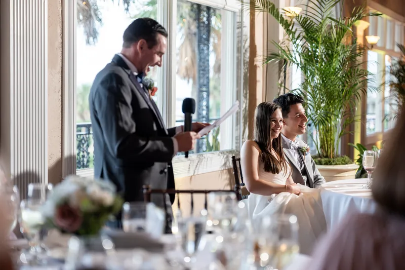 Nathan delivers a toast at The White Room while Sarah sits beside him, smiling as guests listen.
