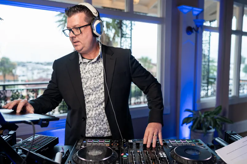 DJ in glasses and blazer operates equipment at The White Room's elevated reception space with waterfront views behind him.