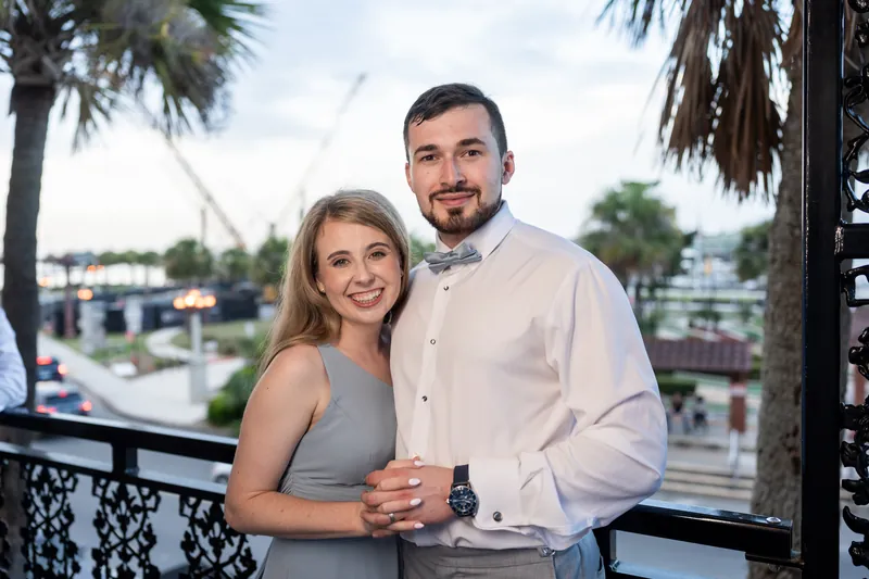 Sarah and Nathan stand together on a balcony overlooking palm trees and waterfront at The White Room.