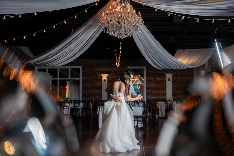 Sarah in flowing white gown stands alone under a crystal chandelier and draped ceiling during The White Room reception.