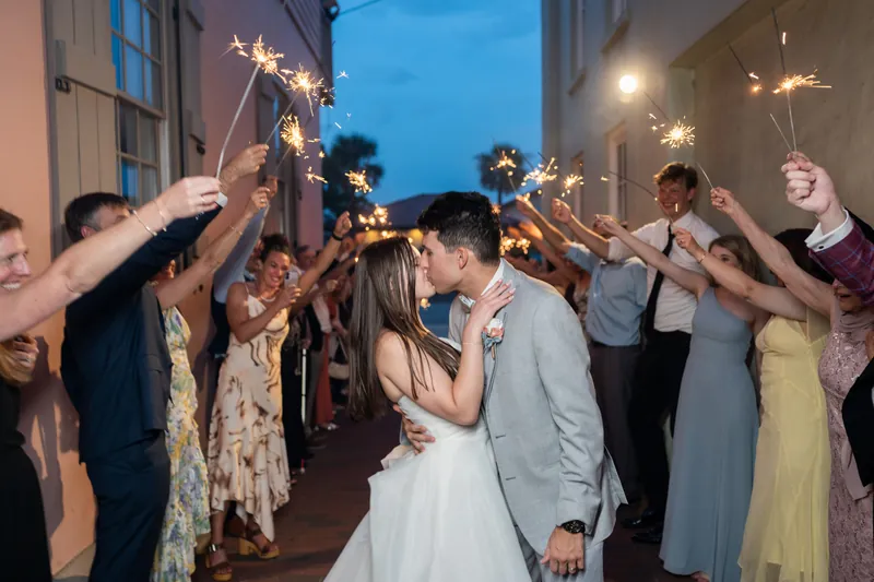 Nathan and Sarah kiss while sparklers light the night sky around them, surrounded by cheering guests.