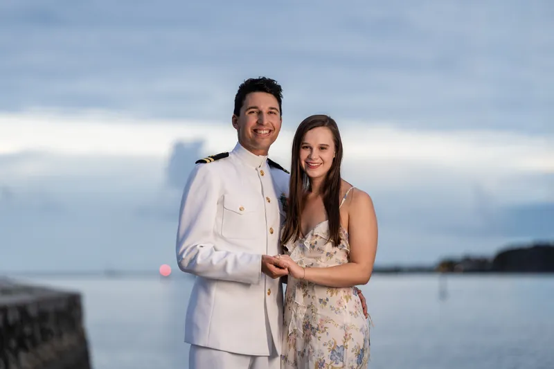 The pair link arms while standing by the bay at Castillo de San Marcos, with moody clouds overhead and water stretching behind them.