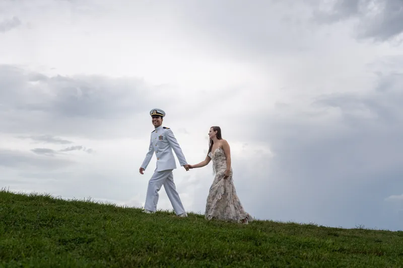 Sarah Katherine and Nathan hold hands walking across a grass slope beneath dramatic cloudy skies at Castillo de San Marcos.