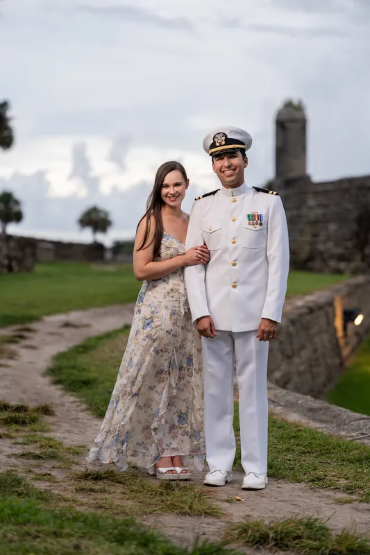Two people in wedding attire stand together on grass with the fort's stone walls behind them under overcast skies.