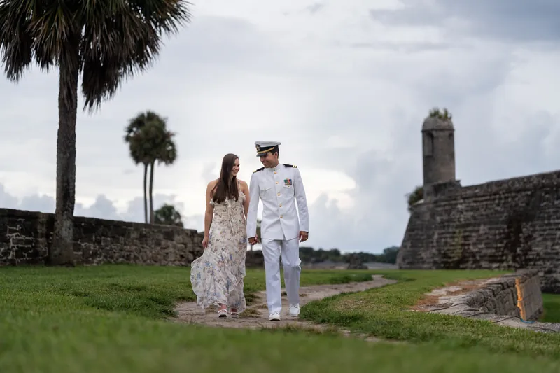 A couple walks across the green grounds with palmetto trees and stone fortifications in the distance.