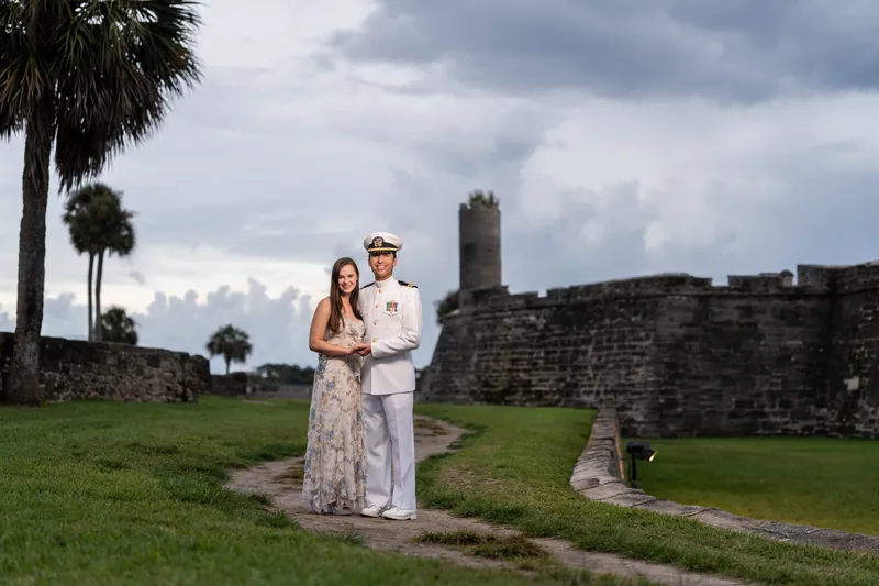 A couple stands together on the lawn at Castillo de San Marcos, the historic stone fortress rising behind them beneath an overcast sky.