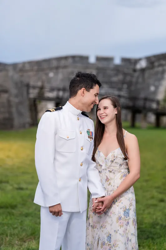 Someone in a white military uniform leans close to a person in a floral gown with the fort's walls softly blurred behind.