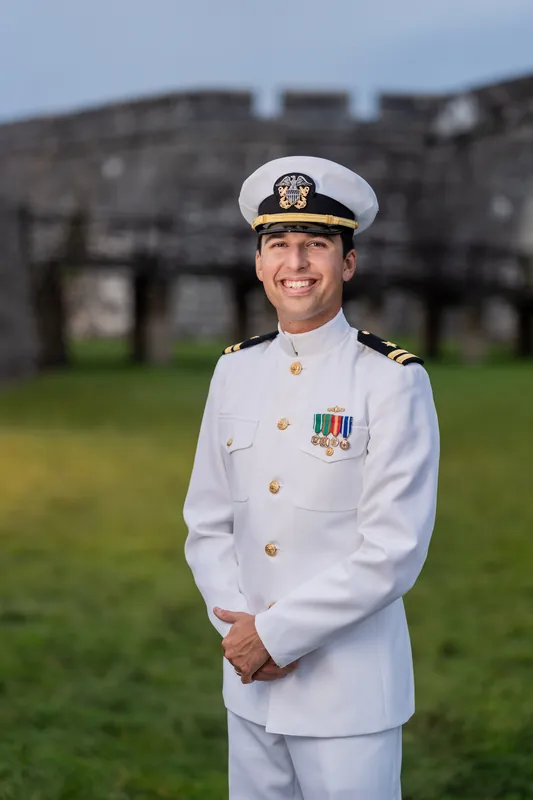 A person in white naval dress uniform with cap and medals stands smiling on the grassy grounds with fortress walls behind.