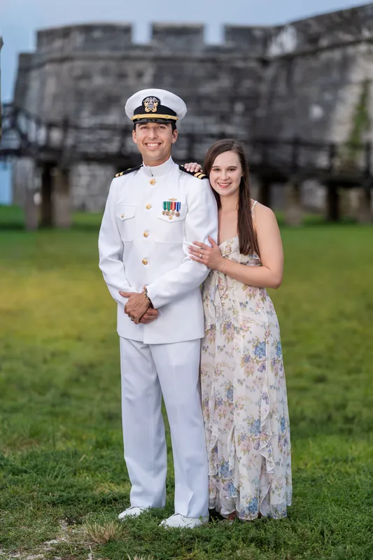 A person in white naval dress uniform and another in a floral gown pose on the grass with the fort's stone bastions visible behind them.