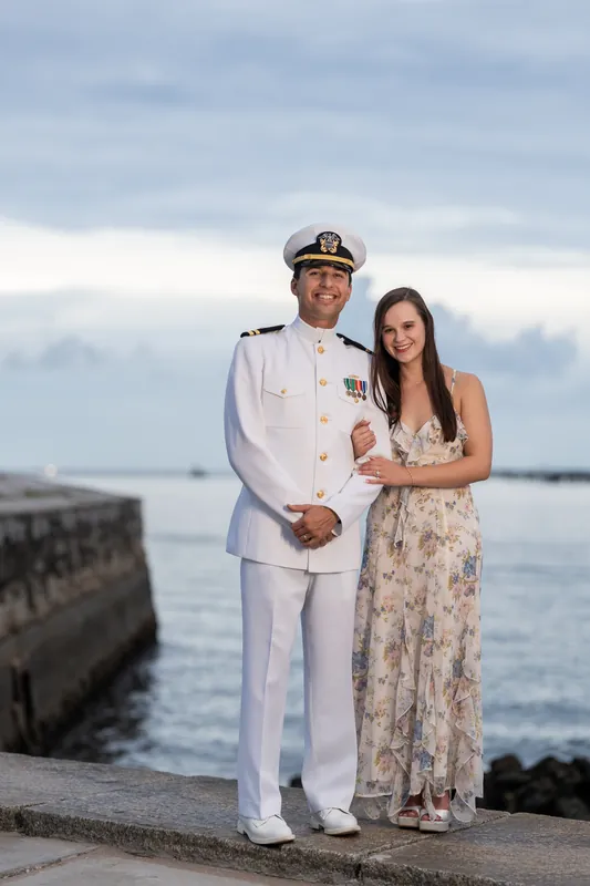 Two people in white dress uniform and floral print stand by the water at Castillo de San Marcos, stone walls behind them.