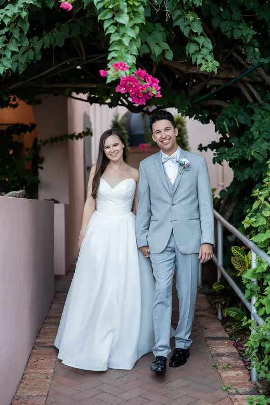 A couple walks hand in hand down a brick pathway lined with vibrant magenta bougainvillea and green foliage.