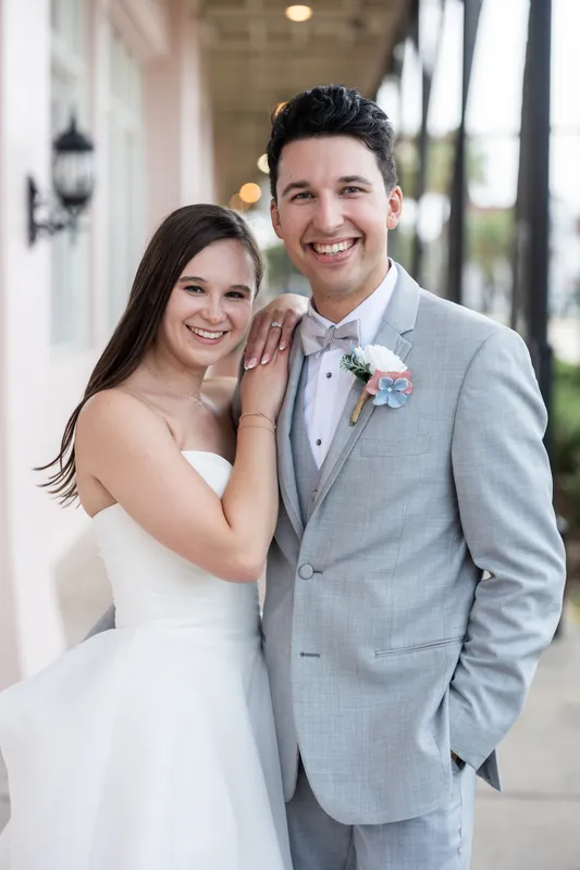 Two people in wedding attire pose together at The White Room, one in a sleeveless white gown and the other in a light gray suit with boutonnière.